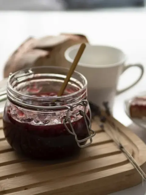 An open jar of dark damson jam with a spoon, beside bread