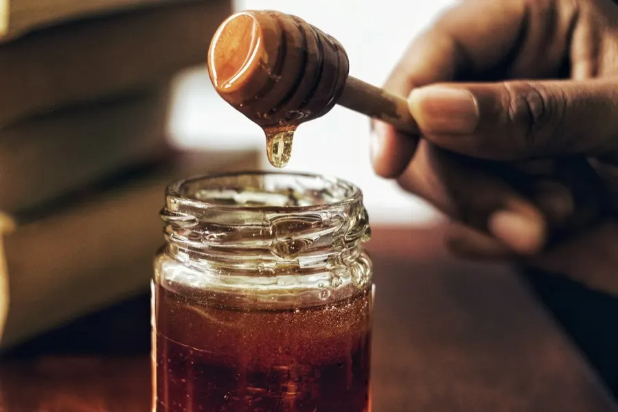 A honey dipper drizzling dark heather honey into an open jar